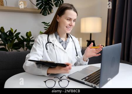 Telehealth concept. Happy attentive general practitioner in uniform and stethoscope using laptop, giving online help, medical support, consultation, t Stock Photo