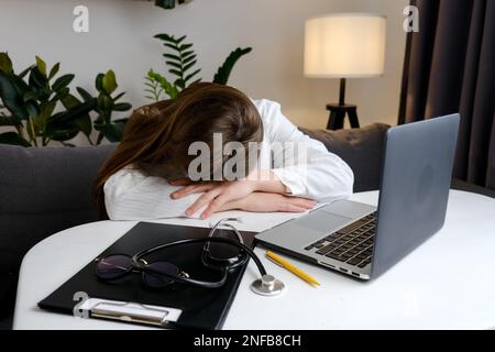 Female doctor in medical uniform sleep in consulting room. Tired exhausted young woman sitting at desk, working on computer with medical documents in Stock Photo