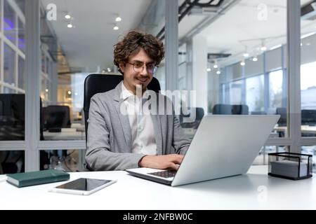 Cheerful and successful businessman inside office working with laptop, bearded man smiling and typing on laptop keyboard, programmer coding software in modern room. Stock Photo