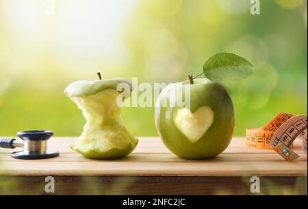 Healthy life and health check concept with eaten apple and whole apple with a bitten heart, stethoscope and tape measure on wooden table with nature b Stock Photo