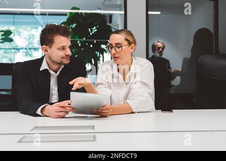 Couple of colleagues man and a woman, sitting by the desk, woman explaining something she has written down on the note pad to her colleague Stock Photo