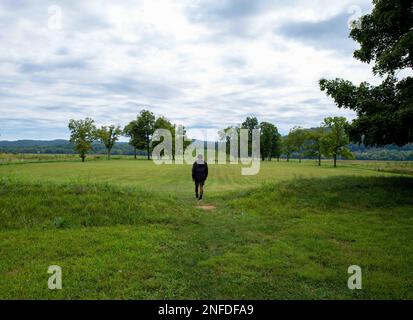 A young man walks toward the Native American Hopewell Culture prehistoric Seip Earthworks burial mound in Ohio. Ancient large long mound. Grass is nea Stock Photo