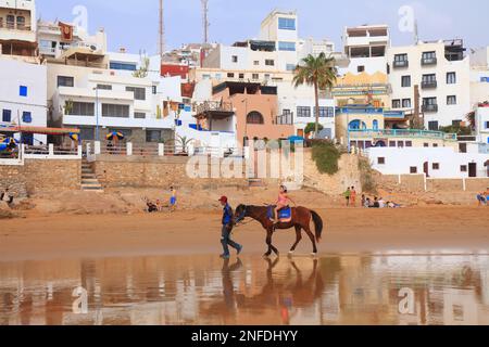 TAGHAZOUT, MOROCCO - FEBRUARY 27, 2022: People visit the beach in Taghazout surfing town near Agadir in Morocco. Stock Photo