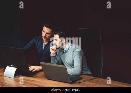 Two business man working in a dark room, on laptop and computer, thinking about ideas for a new campaign  Stock Photo