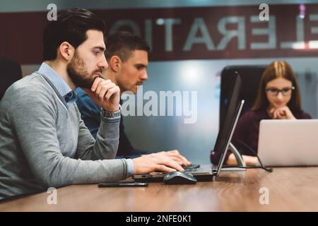 Sitting in front, a young business man working on his laptop, and two of their colleagues, blurred in the background Stock Photo