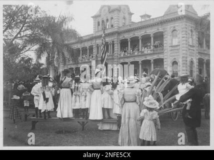 People gathering to see the raising of the American flag and the lowering of the Hawaiian flag at the Executive Builidng (Iolani Palace). Boy on second bench, left, with sailor color is Theodore A. Cooke. People on bench in foreground, L. to r: Alan J. Lowrey, Helen S. Lowrey, Mary Flood, Alice Cooke, Beatrice Castle, Tillie ? . Man of far right is George W. Smith carrying A. Brodie Smith. Stock Photo