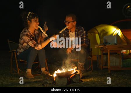 Asian couple roasting marshmallows together at a campfire where they set up a tent to camp by the lake at night. Stock Photo