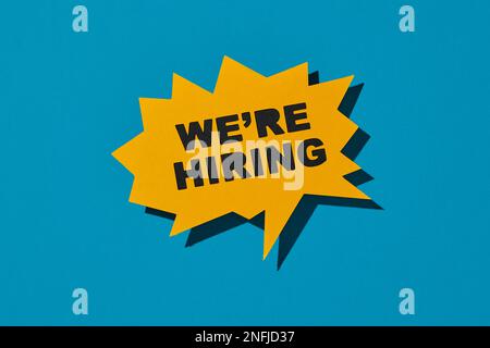 a yellow sign in the shape of a speech bubble reads we are hiring on a blue background Stock Photo