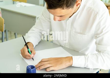 A student writes with a fountain pen while studying at school, sitting at a desk. Portrait of a college guy writing with a pen while completing a task. High quality photo Stock Photo