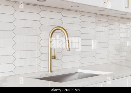 A beautiful kitchen faucet detail with white cabinets, a gold faucet, white marble countertops, and a brown picket ceramic tile backsplash. Stock Photo