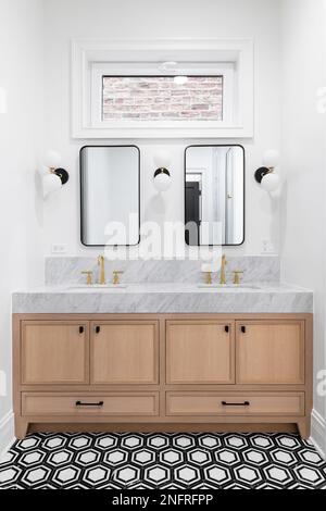 A bathroom with a white oak cabinet, marble countertop, gold faucets, black and white tiled floor, and sconces around black mirrors. Stock Photo