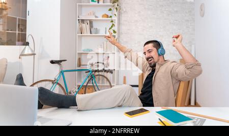 Happy smiling young man in headphones with laptop computer listening music, dancing in home office. Stock Photo