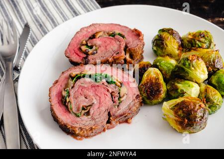Stuffed Flank Steak with Prosciutto and Mushrooms Dinner: Slices of steak roulade served on a plate with roasted Brussels sprouts Stock Photo