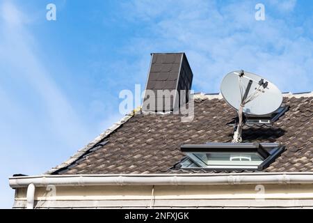 A fragment of a tiled roof of a house with chimney, satellite dish and window against a blue sky with light clouds. Close up. Stock Photo