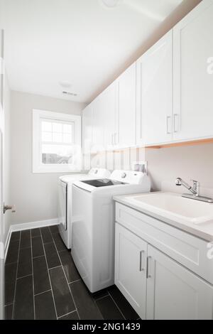 A laundry room with dark grey tiled floor, white cabinets and appliances. Stock Photo