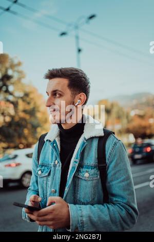 A student with a backpack using a smartphone on the street, checking email, writing messages and listening to music on headphones. Stock Photo