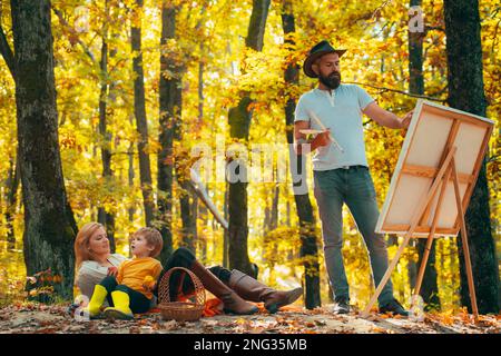 Handsome bearded man draws a picture of his young family in the park using a palette with paints and a spatula. Drawing easel and canvas with a Stock Photo