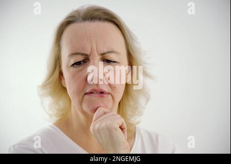 Photo of thoughtful woman bites lower lip, looks with dreamy expression aside, wearing stylish beige hoodie, stands against white wall. Copy space for your text. High quality photo Stock Photo