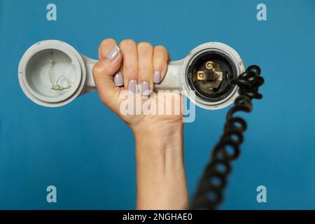 female hand holding an old broken telephone receiver on a wire on a blue isolated background, the telephone receiver on the prododi is broken Stock Photo