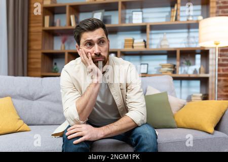 Overworked sad man sitting alone on sofa, mature businessman at home holding hands on head in depression in living room in casual clothes. Stock Photo