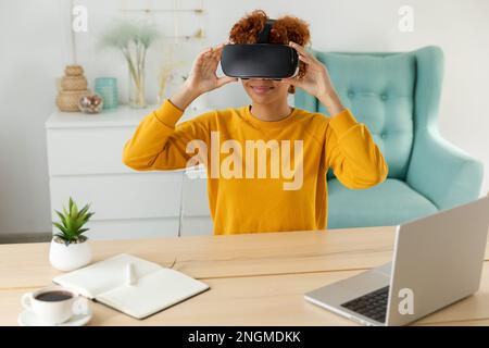 African american young woman wearing using virtual reality metaverse VR glasses headset at home. Girl puts on virtual reality helmet. Technology simulation hi-tech videogame concept Stock Photo