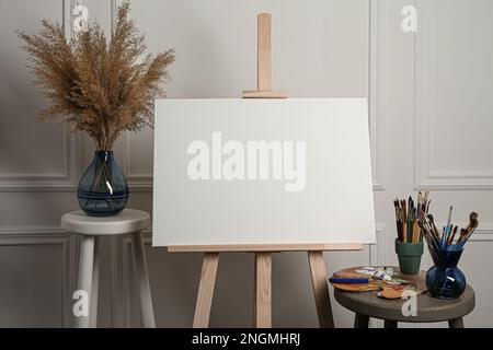 Easel with blank canvas, brushes, paints and palette in studio Stock Photo