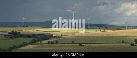 Landscape in early summer with sun breaking through dark clouds and highlighting fields with tractors working and wind turbines in near distance Stock Photo
