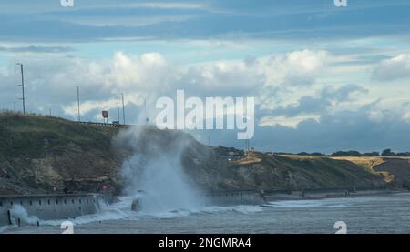 Heavy waves crash onto the promenade at Seaham throwing up columns of spray with cliffs and coastal road caught in the sunlight Stock Photo