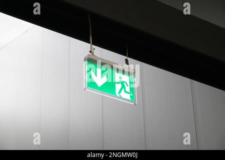 emergency exit or fire escape sign with green running man symbol and directional arrow hanging from ceiling Stock Photo