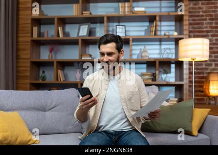Cheerful and smiling man at home paying bills and checks, using banking app on phone sitting on sofa in living room holding bills. Stock Photo