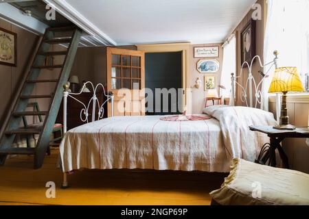 Double bed with white painted wrought iron headboard and footboard with brass ball finials in upstairs guest bedroom with wooden miller's stairs. Stock Photo