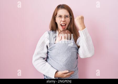 Young caucasian woman holding and carrying baby on a sling annoyed and frustrated shouting with anger, yelling crazy with anger and hand raised Stock Photo