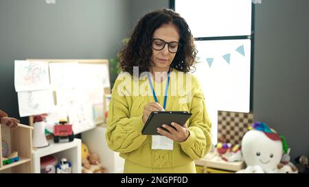 Middle age hispanic woman preschool teacher using touchpad at kindergarten Stock Photo