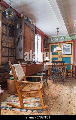 Antique rocking chair, cast iron pan with long handle hanging from piece sur piece log wall, wooden cabinets and pinewood dining table in old house. Stock Photo