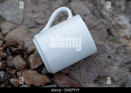 White mug, mock up, empty space for artwork, text, standing on stone, outdoors, green grass, trees, sky in the background, soft sunlight, nature Stock Photo