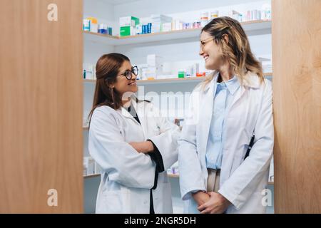 Two female pharmacists standing in a drug store and smiling at each other. Happy healthcare professionals working together in a pharmacy. Stock Photo
