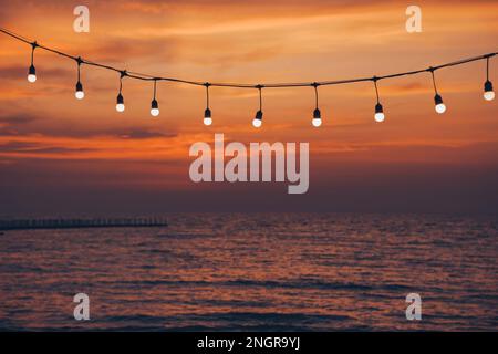 light bulbs on string wire against sunset sky by the beach Stock Photo