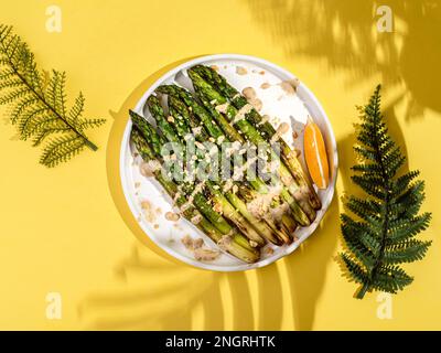 cooked asparagus in a plate. asparagus fried with olive oil, drizzled with sauce and sprinkled with nuts, next to a slice of lemon. On a yellow backgr Stock Photo