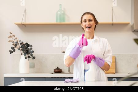 Attractive housewife holds a spray bottle with cleaning agent in her hand and shows a thumbs up.  Stock Photo