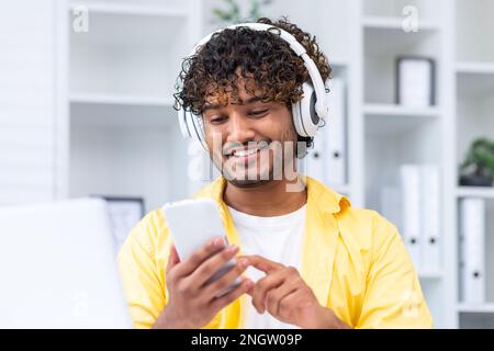 Young smiling hispanic man at home close-up, man in headphones listening to music online books and audio podcasts, using phone typing messages and watching videos, holding smartphone. Stock Photo