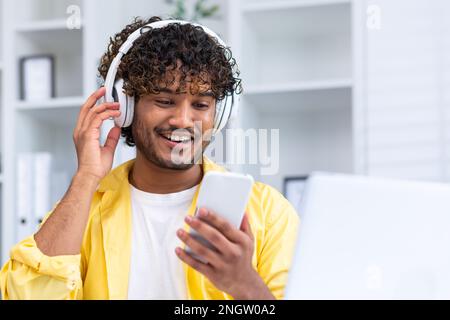 Happy smiling indian man watching video on smartphone close up in yellow shirt in home office, man smiling and surfing online stream using headphones. Stock Photo