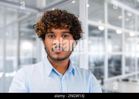 Close-up portrait of young successful hispanic businessman inside office, man in blue shirt concentrating and thinking looking at camera, programmer at workplace with curly hair. Stock Photo