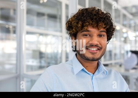 Young smiling indian programmer close up smiling and looking at camera, portrait of man with curly hair and blue shirt inside office at work, businessman entrepreneur with beard working on project. Stock Photo