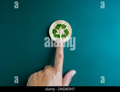 Green recycle icon symbol on round wooden block, pointing by hand isolated on blue background. Ecology, sustainability cooperation, reduce, reuse and Stock Photo