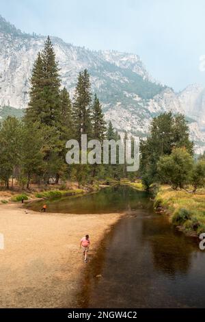 View of the Merced River and mountains with smoke from the fires in the background. A man and a couple of children walk along the river Stock Photo