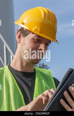Male engineer with short brown hair, wearing yellow hard hat and reflective vest, inspecting wind turbine using digital tablet, smiling, close-up Stock Photo