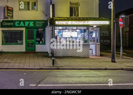 Chinese takeaway at night in a suburban high street with customers waiting inside. Next to a traditional British Cafe. Cultural Contrast. Stock Photo