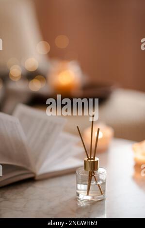 Home liquid fragrance in glass bottle and bamboo sticks over paper book on marble table in living room close up. Cozy atmosphere. Stock Photo