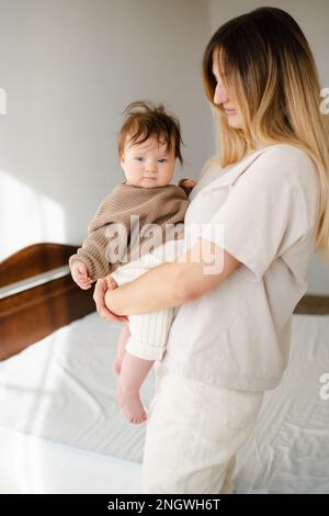 Cute infant baby girl under 1 year old on mother hands wear casual knit sweater and pants in bedroom. Look at camera. Motherhood. Stock Photo