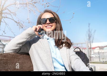 Young brunette woman talks on a cell phone and smiles wile sitting on bench outdoors. Stock Photo